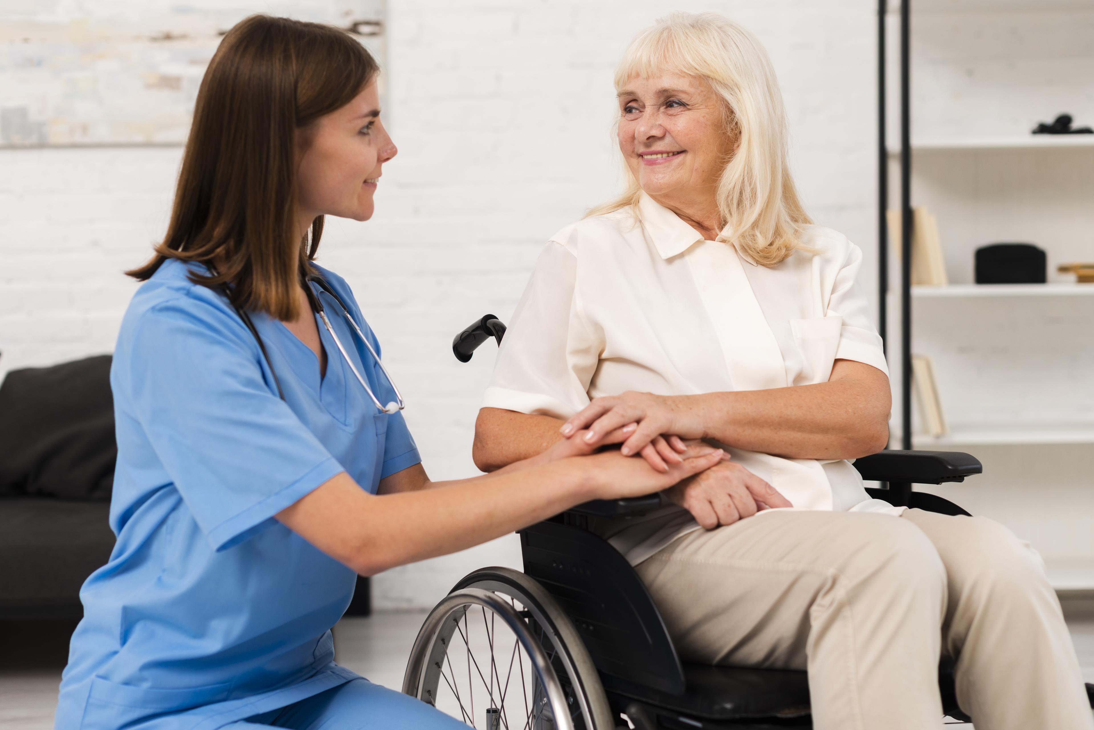 A female nurse kneeling down beside an old woman on a wheel chair.